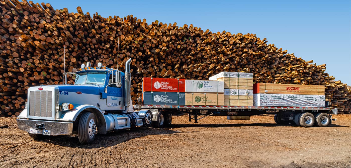 A photo of a South Coast Lumber Company truck loaded with products in front of a tall stack of logs